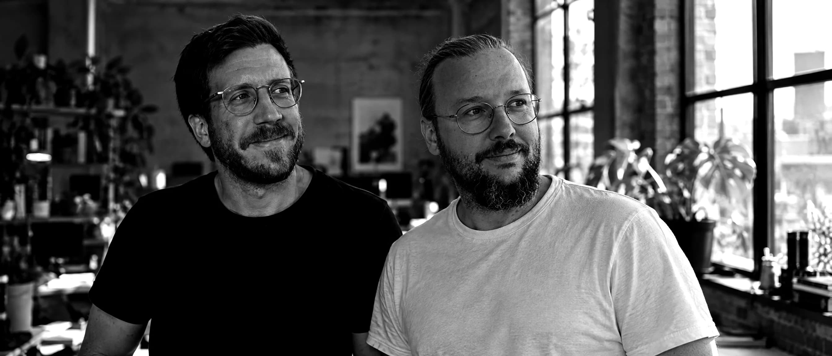 Schwarzweiß-Aufnahme von Tobias Vonau und Philipp Russner in einem Büro mit Backsteinwand, hohen Fenstern und Pflanzen.